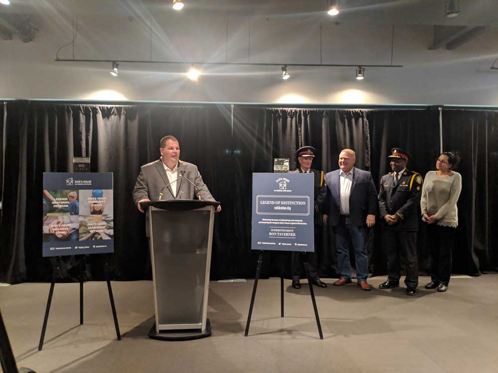 man in grey suit standing at podium during jake's house event with police officers woman and doug ford in the background