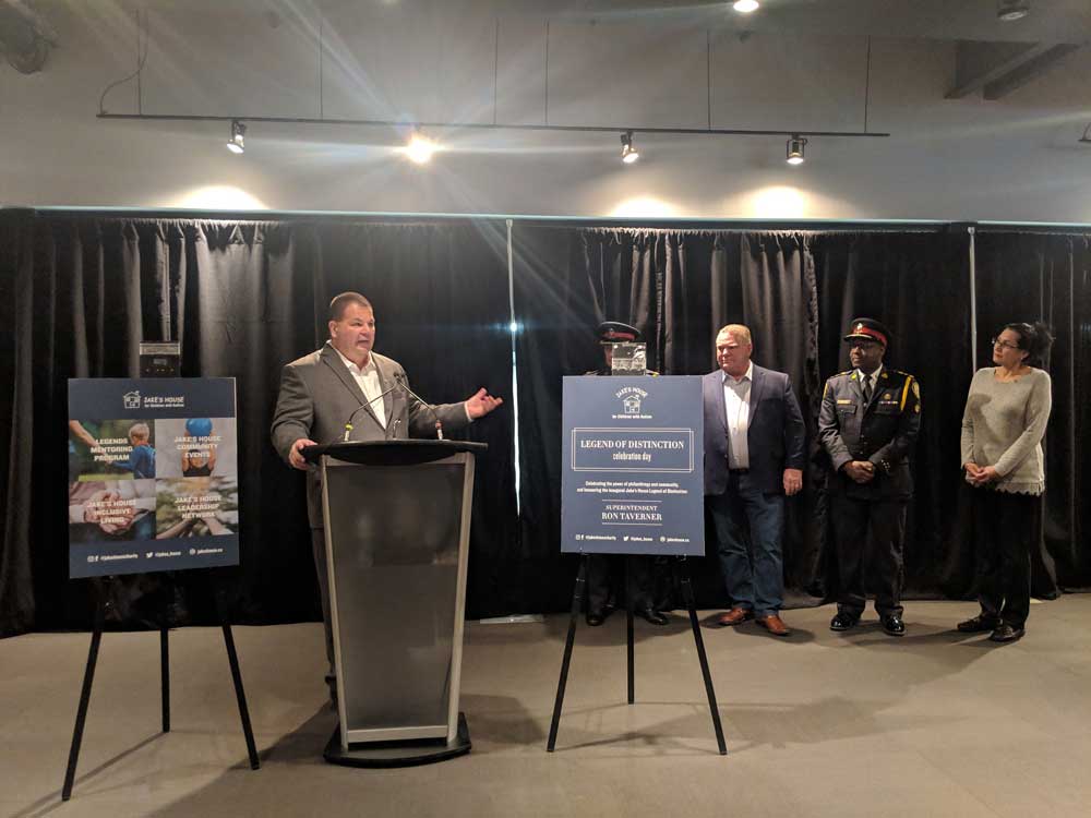 man in grey suit standing at podium during jake's house event with police officers woman and doug ford in the background