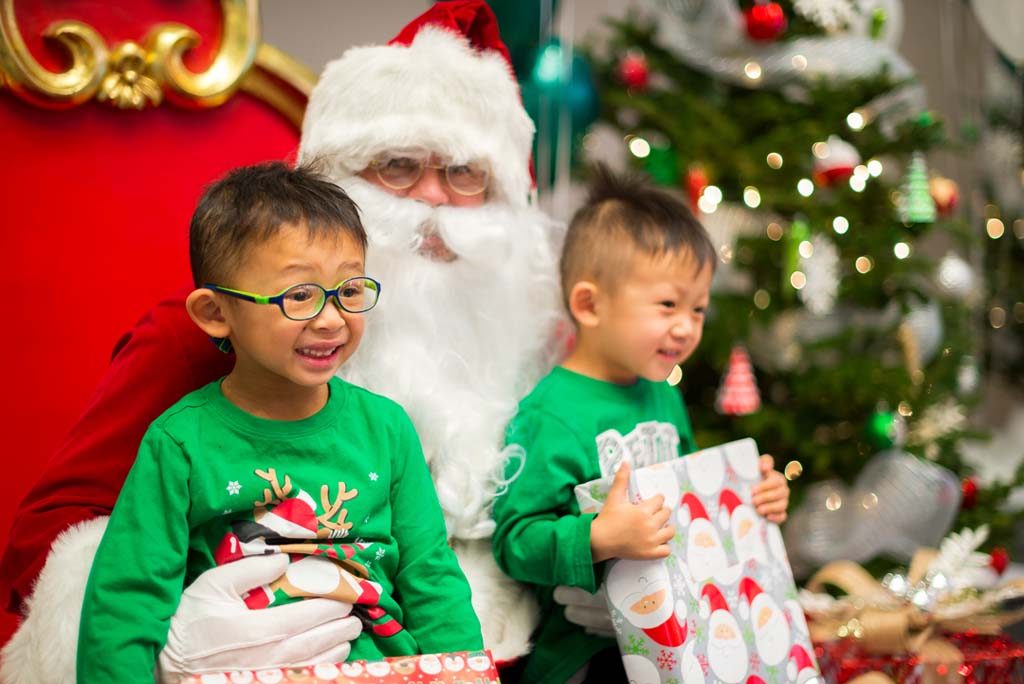Two smiling children in green sweaters sitting on Santa's knee and holding wrapped presents