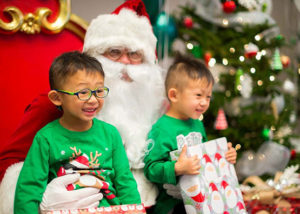 Two smiling children in green sweaters sitting on Santa's knee and holding wrapped presents