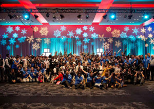 A group photo at a Holiday party lit by red and blue lights, red balloons, and snow flake projections.