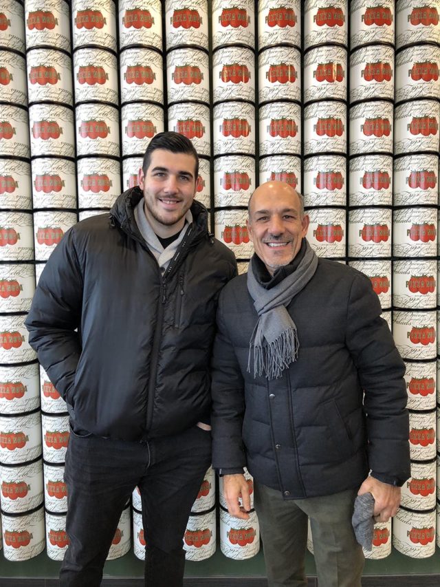 two men posing in front of a wall of stacked canned tomatoes