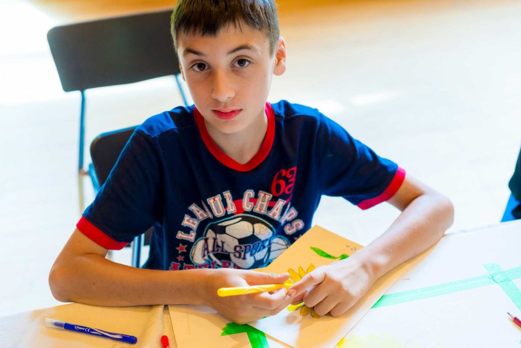 boy sitting at table in dark blue shirt holding marker over artwork