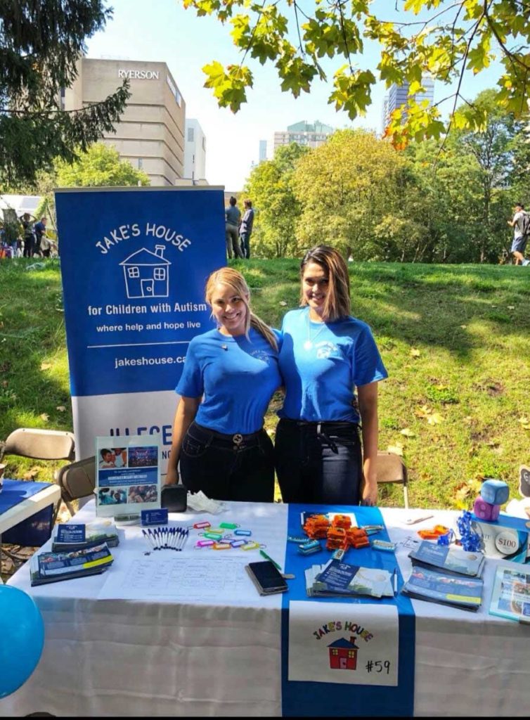 two jake's house volunteers wearing blue shirts standing behind table with banner