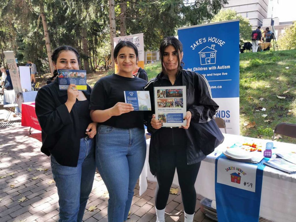 three women holding jake's house flyers in front of table outside