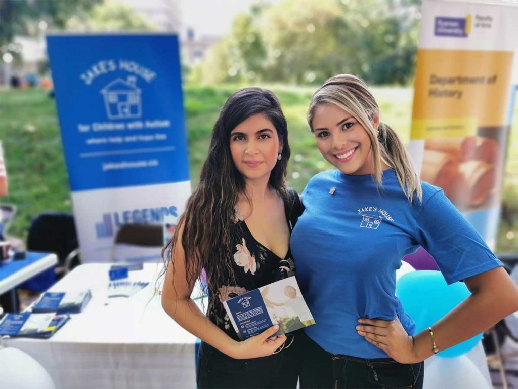 two women posing in front of jake's house volunteer table